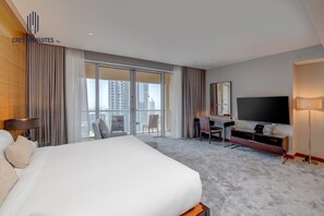 Ein Hotelzimmer mit einem großen Bett, einem Schreibtisch, einem Sessel, einem Fernseher und einem Balkon mit Blick auf die Stadt.