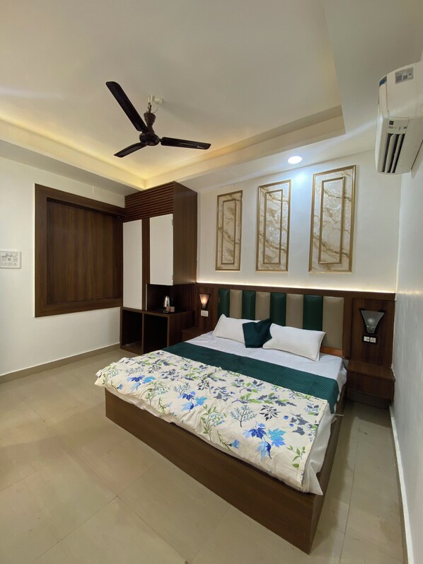 A bedroom with a bed, a ceiling fan, wall-mounted artwork, and a bedside table. - HOTEL RMG & GARDEN (Navi Mumbai)