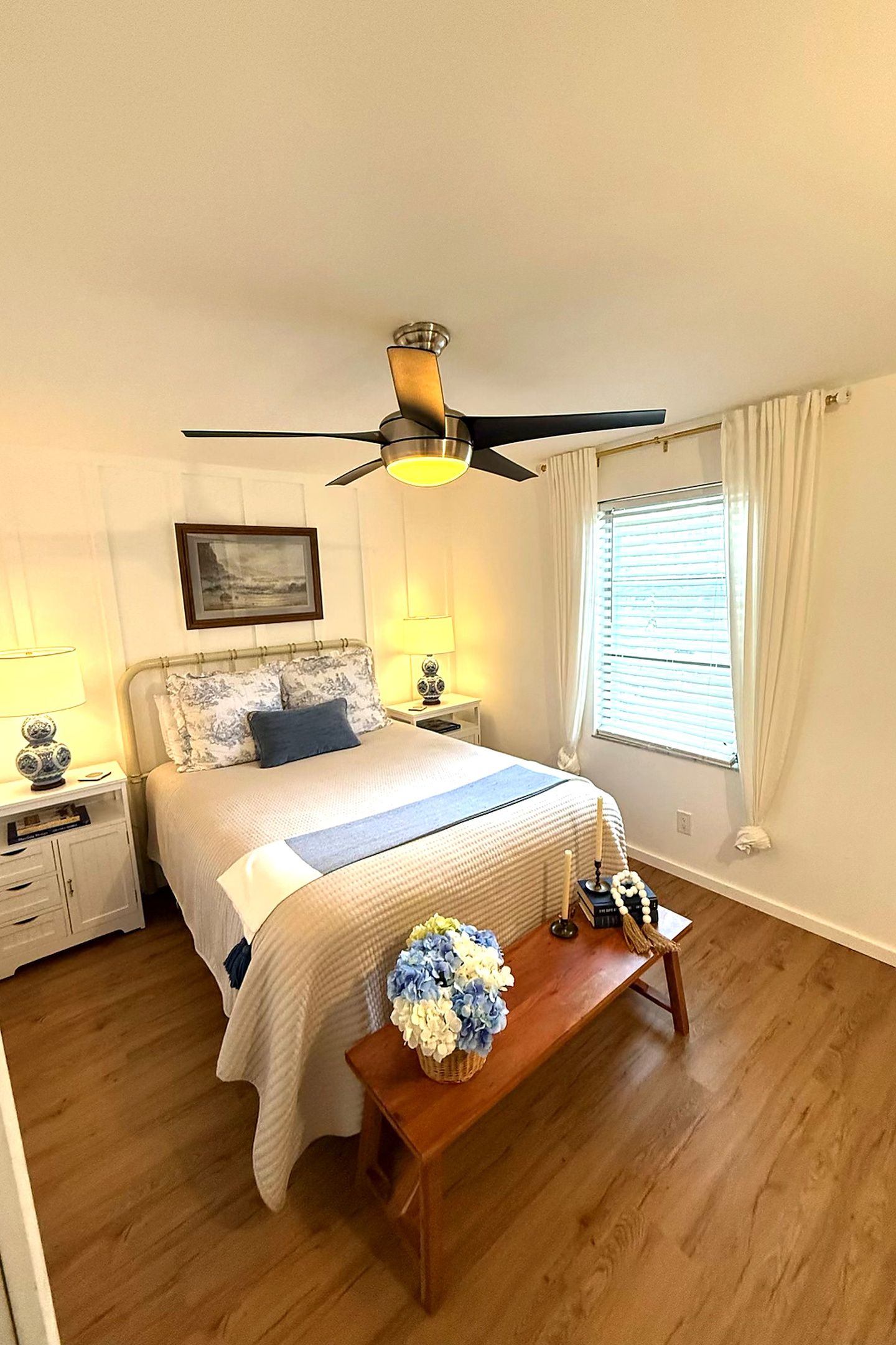 A bedroom with a bed, bedside lamps, a ceiling fan, a window with curtains, and a wooden bench with a vase of flowers.