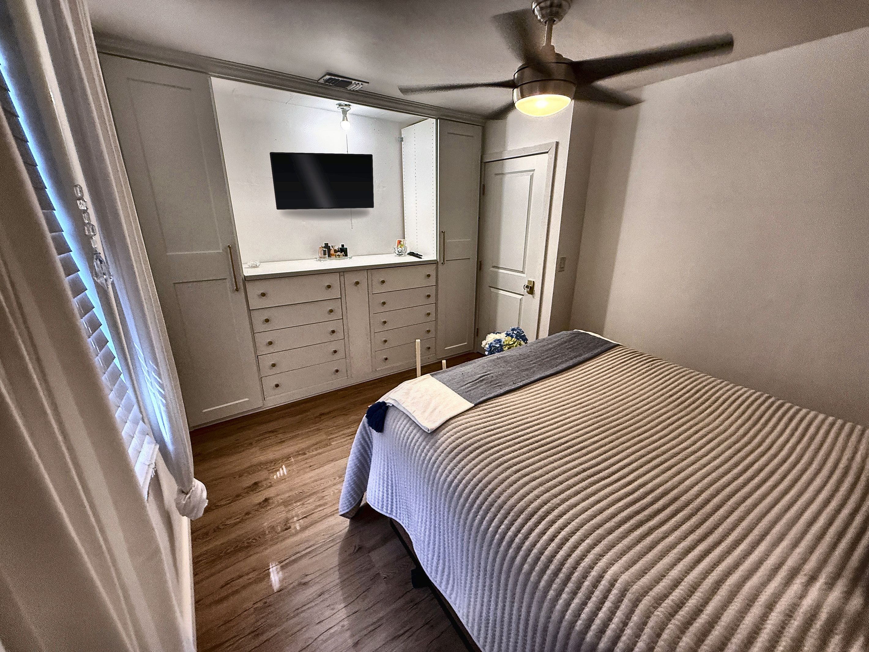 Une chambre à coucher avec un lit, un ventilateur de plafond, un téléviseur fixé au mur et une commode.
