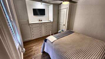 Une chambre à coucher avec un lit, un ventilateur de plafond, un téléviseur fixé au mur et une commode.