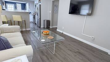 Una sala moderna con una mesa de centro de vidrio, un televisor de pantalla plana montado en la pared y una cocina al fondo.