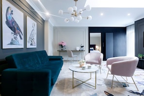 Una sala de estar moderna con un sofá color turquesa, un sillón rosa y una mesa de centro. Hay obras de arte enmarcadas en la pared, una araña y una vista a otra habitación.