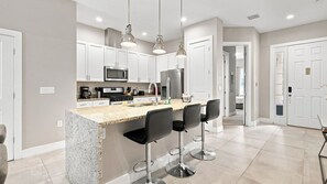 A modern kitchen with white cabinets, stainless steel appliances, and a granite countertop. There are three bar stools and pendant lights above the counter. - Summerville Resort Prime Location Near Disney (Kissimmee)
