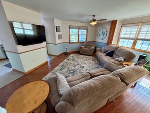 A living room with a sectional sofa, a flat-screen TV, a ceiling fan, and large windows. - Ultimate Family Shore Getaway | 5BR Townhome Sleeps 12 in North Wildwood (North Wildwood)
