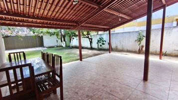 Un patio techado con mesa y sillas, piso de baldosas y vista a un jardín cubierto de césped y árboles.