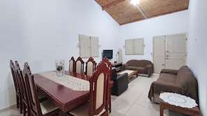 Un comedor con mesa y sillas de madera, televisión y sofá en una habitación con paredes blancas y techo de madera.
