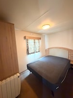 Acomodação pequena e individual, com uma cama, radiador, janela com cortinas e luminária no teto.