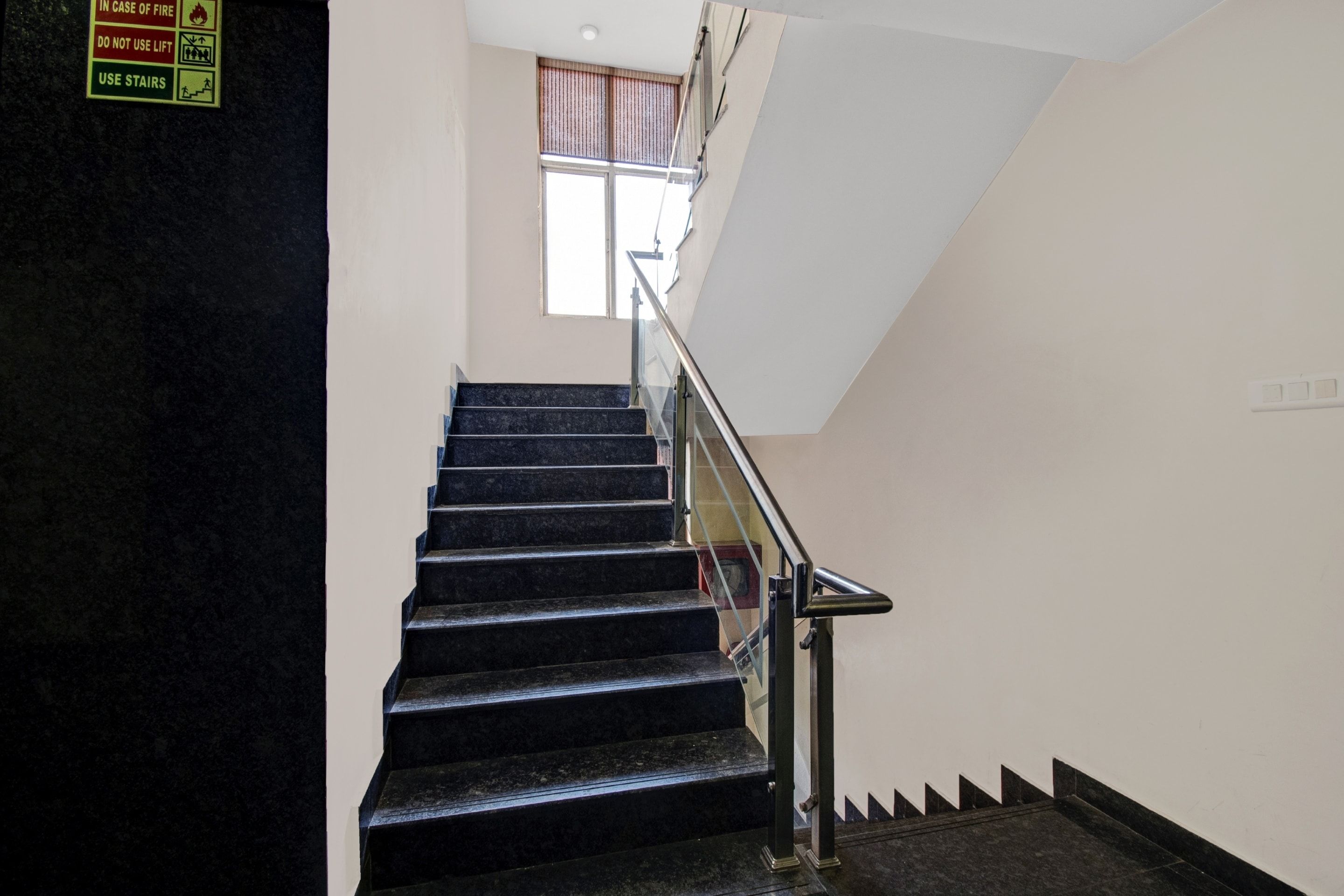 Una escalera con escalones negros y barandilla metálica, que conduce a un rellano con ventana.