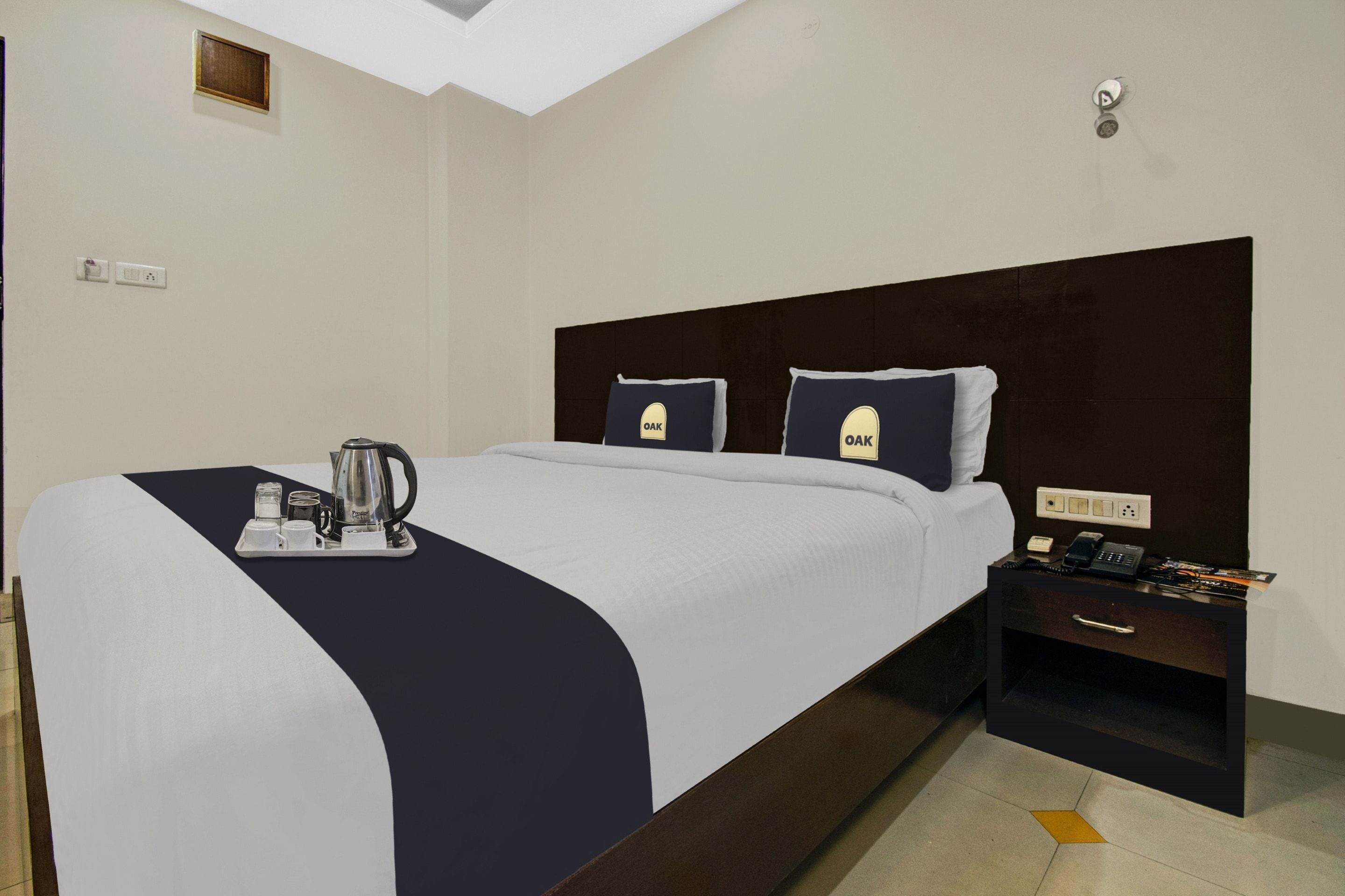 Una habitación de hotel con cama, una mesita de noche con teléfono, una bandeja con tetera y un interruptor de luz en la pared.