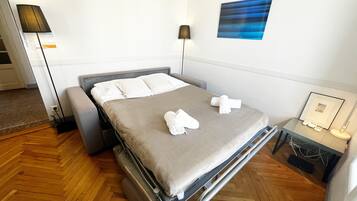 Een netjes opgemaakt bed met een grijs matras en witte lakens, een nachtkastje met een lamp, en een aan de muur bevestigd kunstwerk.
