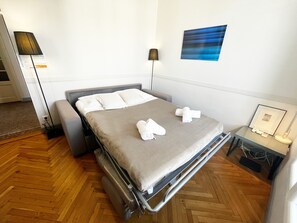 Een netjes opgemaakt bed met een grijs matras en witte lakens, een nachtkastje met een lamp, en een aan de muur bevestigd kunstwerk.