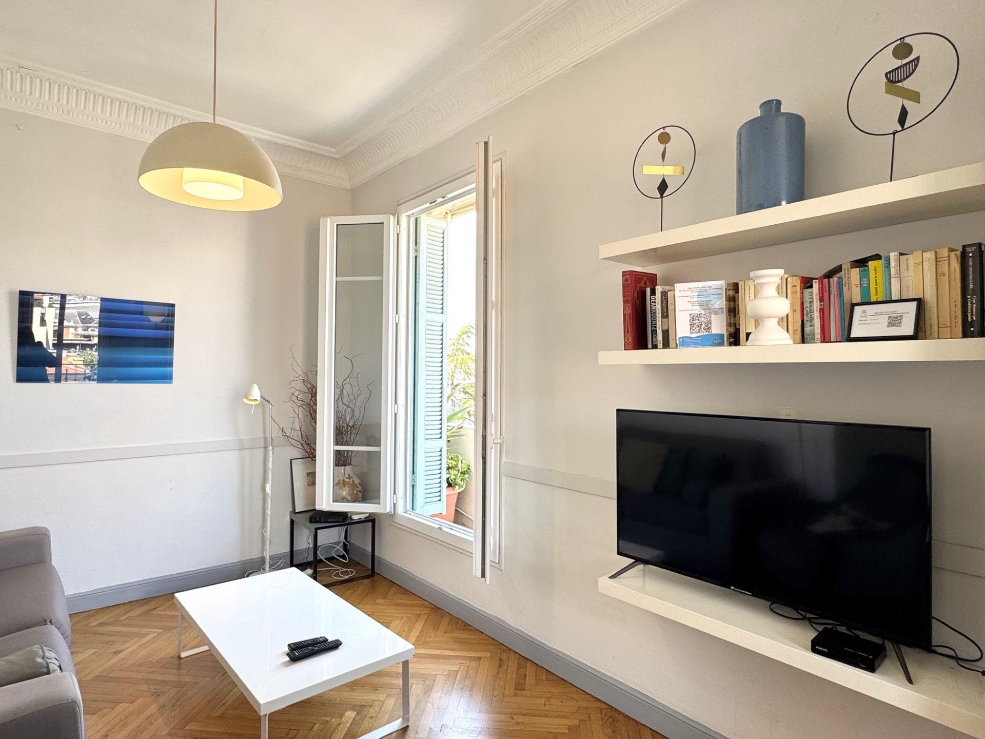 Un salon comprenant un téléviseur à écran plat, une table basse blanche, un canapé gris, un lampadaire et une bibliothèque remplie de livres et d’objets décoratifs.