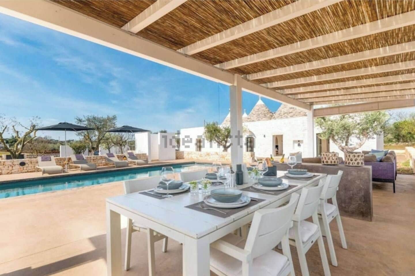 An outdoor dining area with a pool, white furniture, and a covered section with a thatched roof.