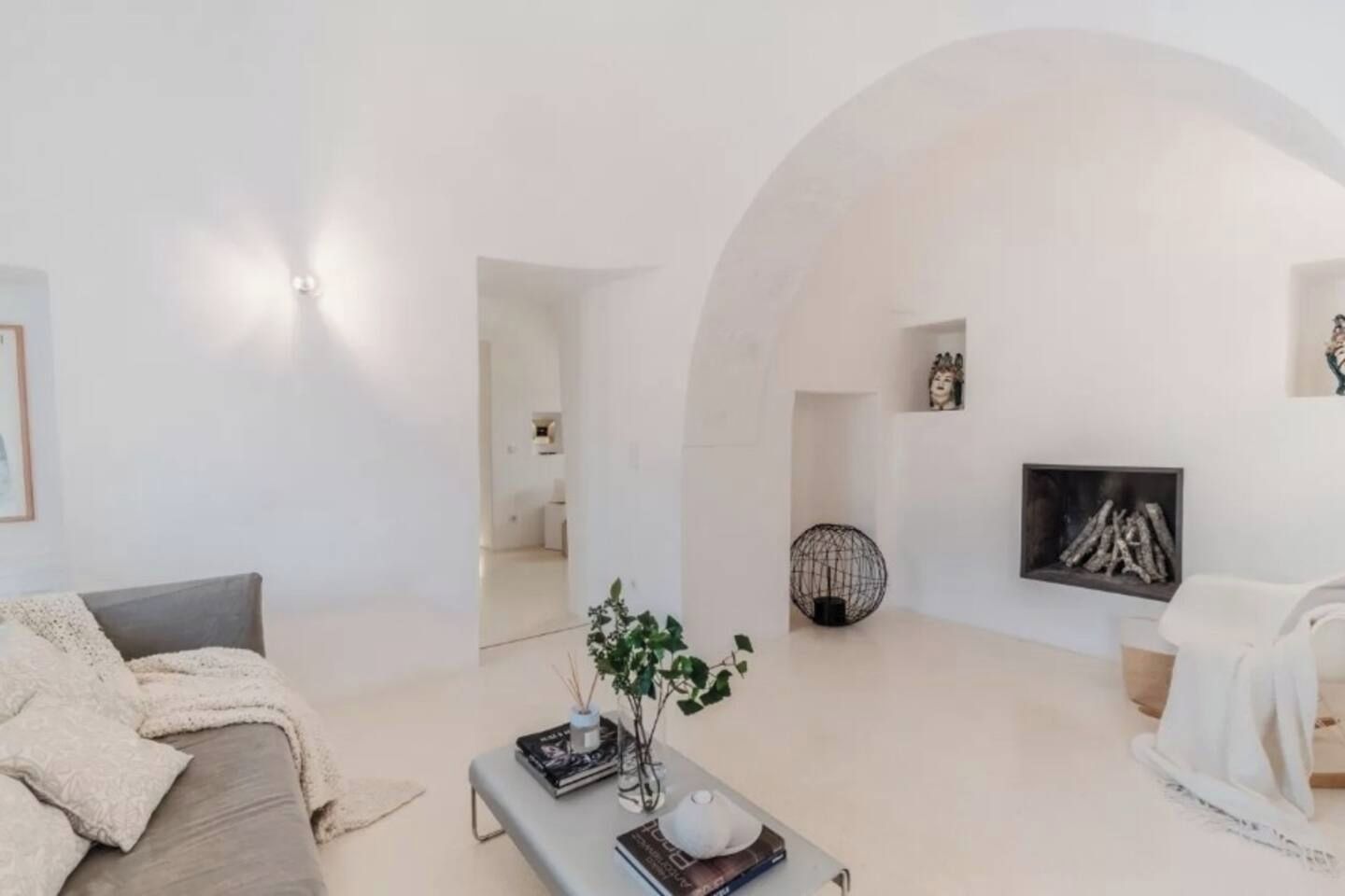 Un salon moderne comprenant un canapé, une table basse avec des livres et un vase, une cheminée ornée d’un tableau encadré, et une porte en arc donnant sur une autre pièce.