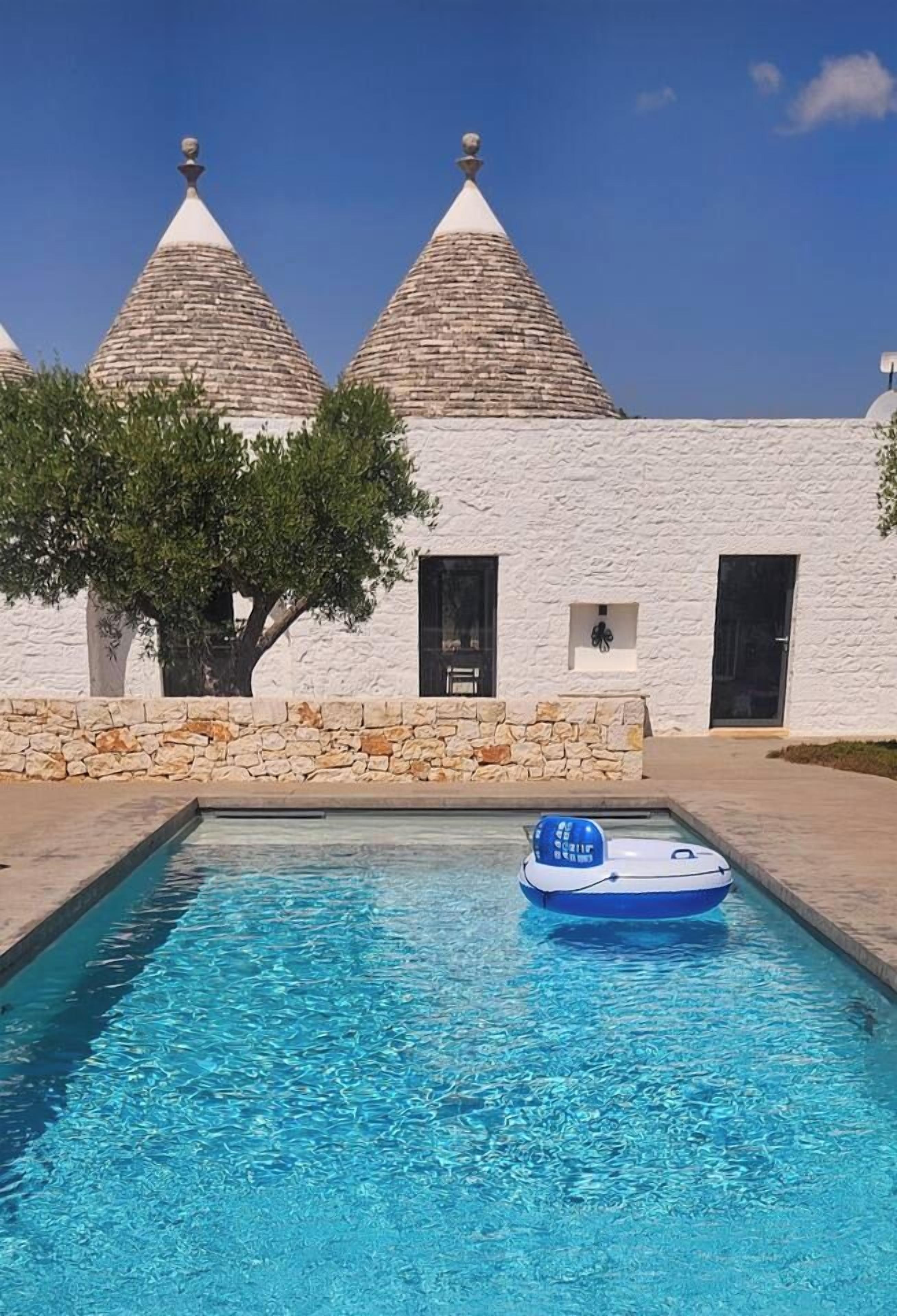 Piscina com um flutuador azul inflável no centro, um edifício branco com paredes de pedra e dois telhados cônicos, e uma árvore em frente ao edifício.