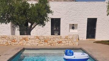 Piscina com um flutuador azul inflável no centro, um edifício branco com paredes de pedra e dois telhados cônicos, e uma árvore em frente ao edifício.
