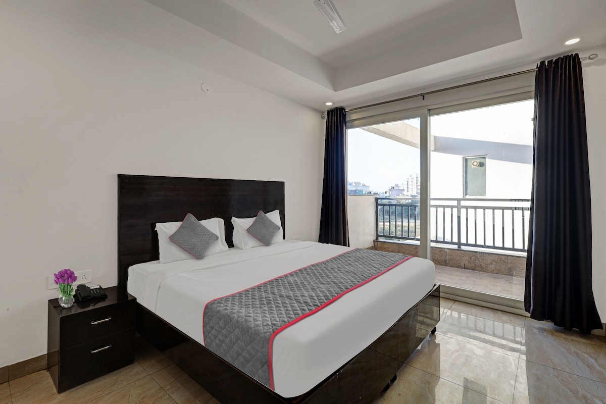 Habitación de hotel con cama, mesita de noche, un jarrón con flores y vistas a la ciudad a través de amplias ventanas.