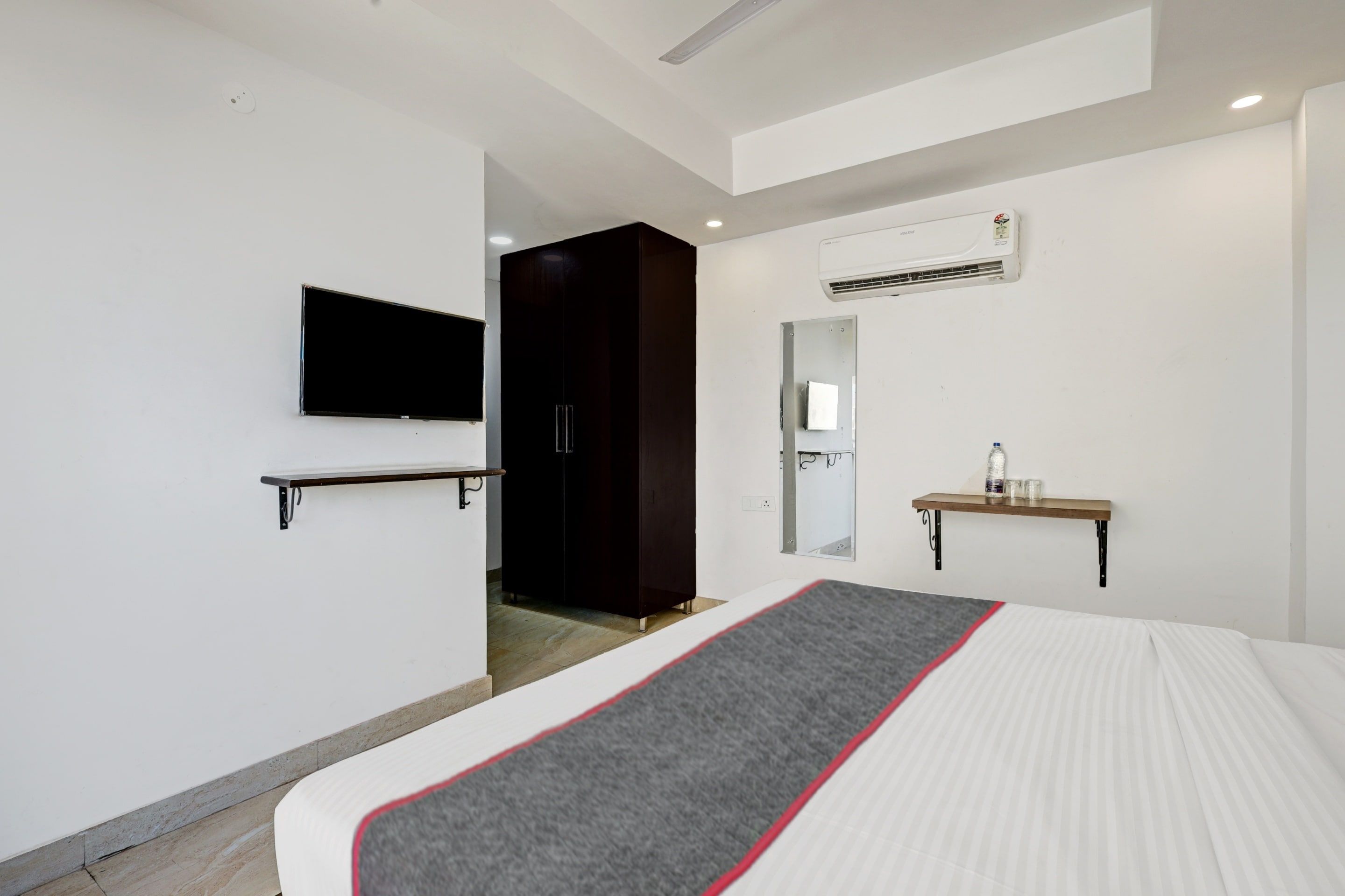 A hotel room with a bed, a TV mounted on the wall, a wardrobe, a mirror, and a small shelf with a bottle and glasses.