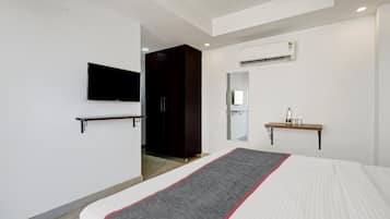 A hotel room with a bed, a TV mounted on the wall, a wardrobe, a mirror, and a small shelf with a bottle and glasses.
