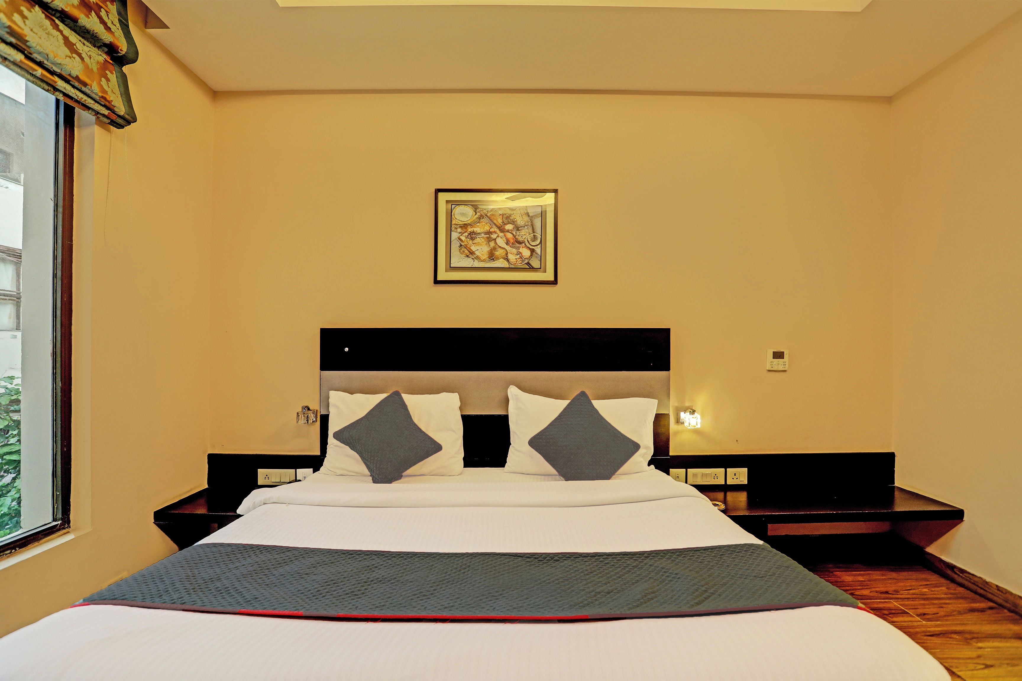 Une chambre d’hôtel comprenant un lit, deux tables de chevet, une petite table, une œuvre d’art accrochée au mur et une fenêtre avec des rideaux.