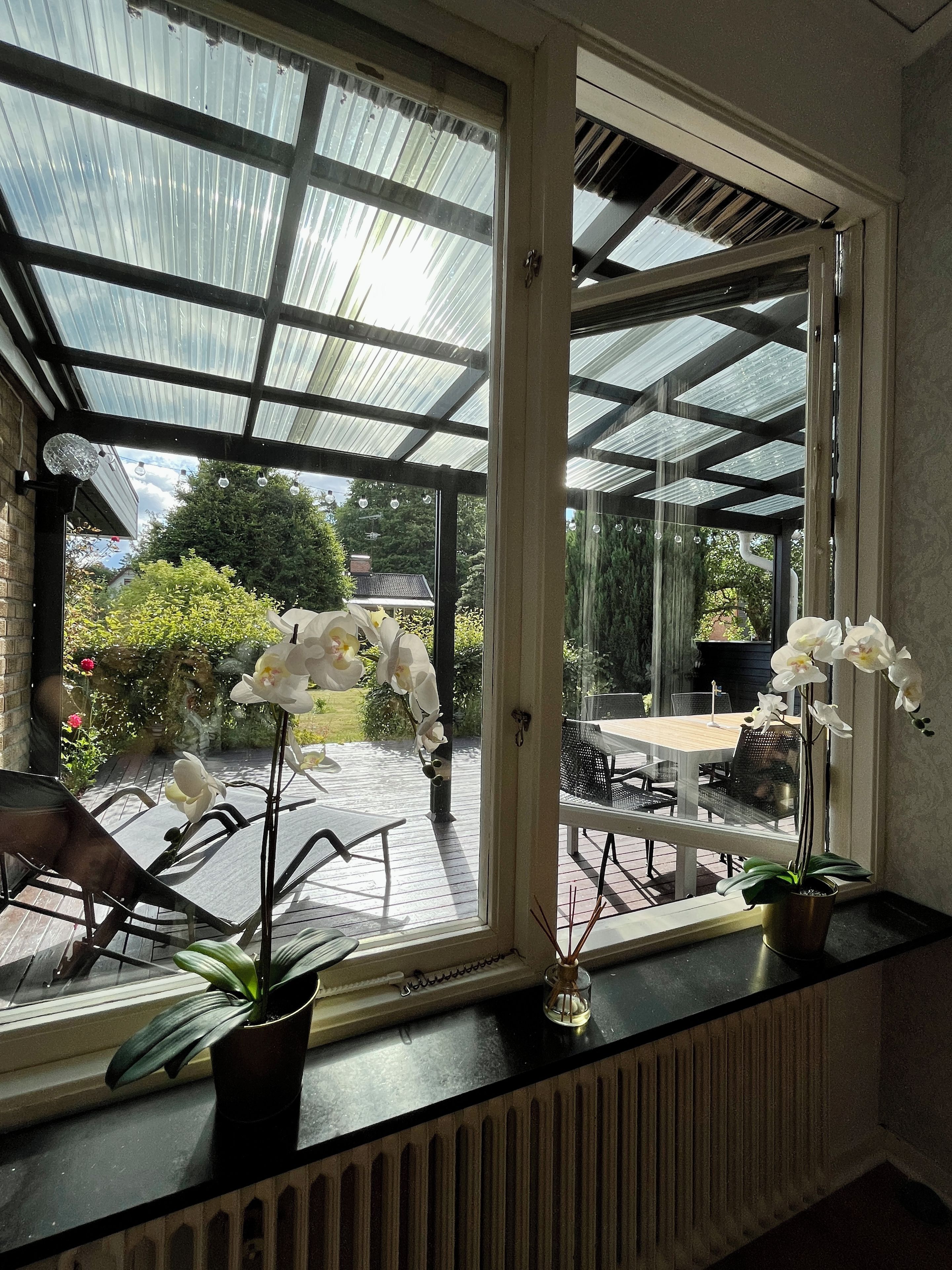 Une véranda avec un toit en verre, une table, des chaises et des plantes en pot.
