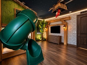 A room with a slide, a TV, a wooden cabinet, and a stone wall. - Ever After Estate | Luxury 12 Bed Home | 9 mins to Disney World (Kissimmee)