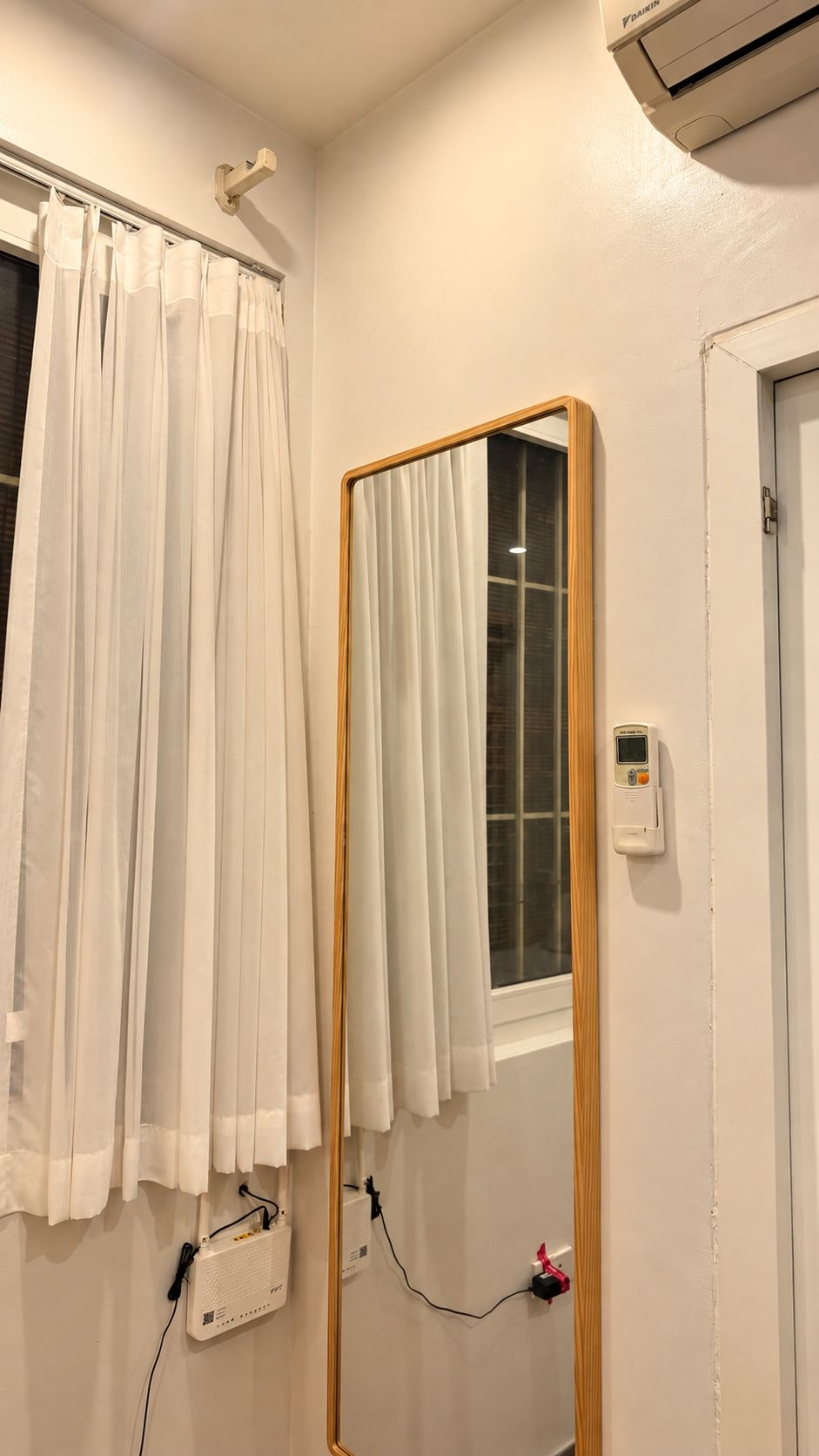 A bathroom with a mirror, a window with curtains, an air conditioning unit, and a wall-mounted device.