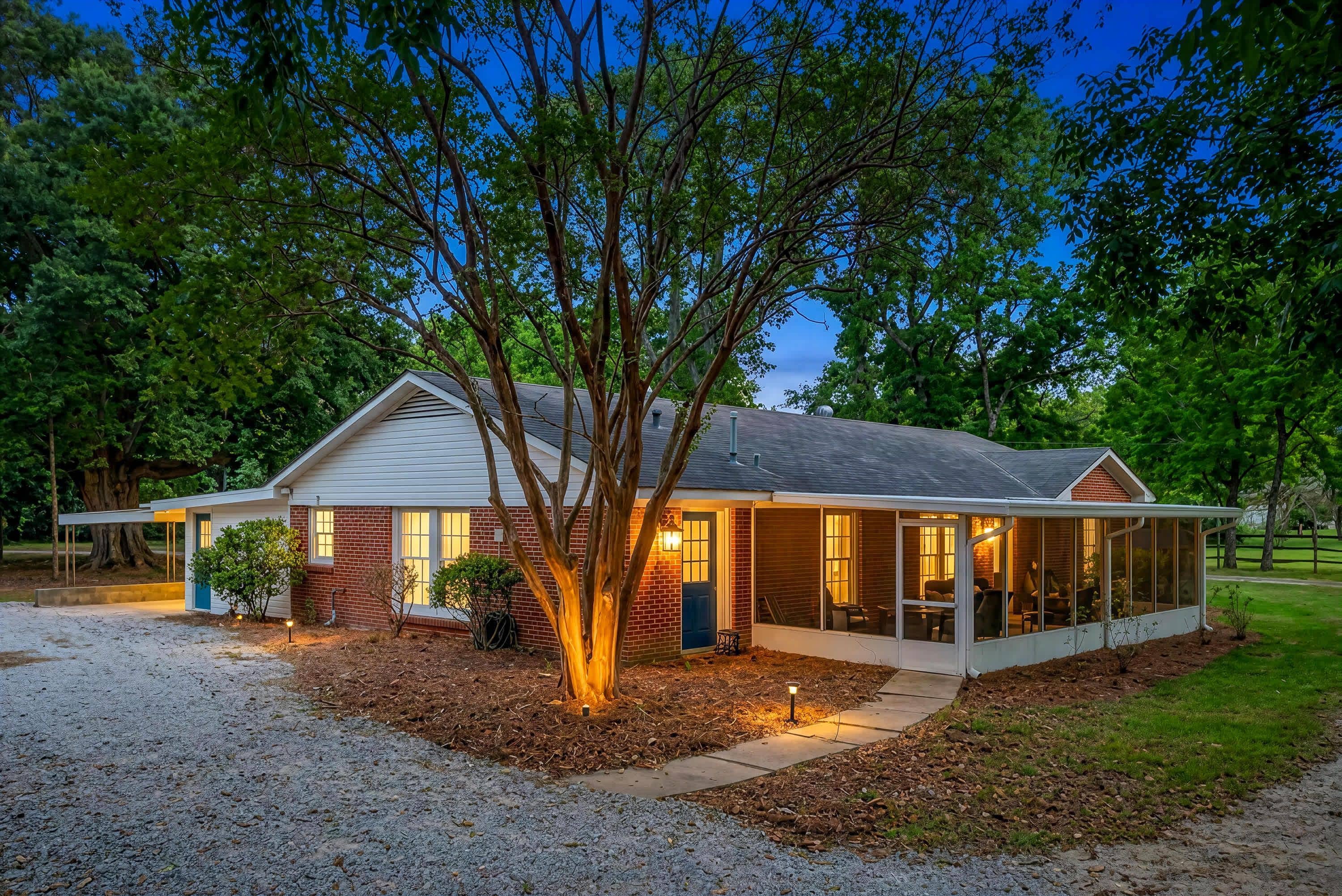 A single-story house with a covered porch, a large tree in front, and a gravel driveway.