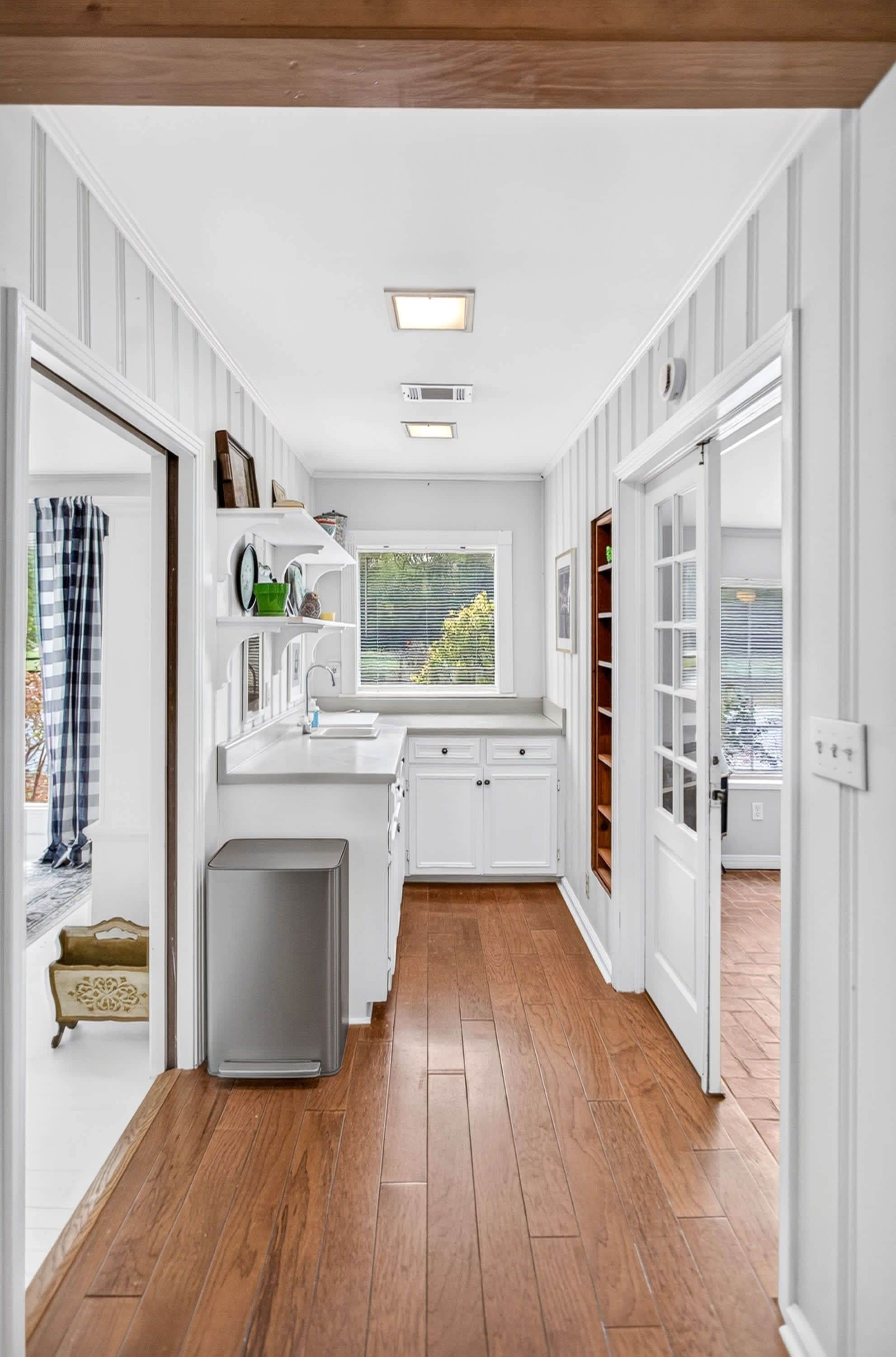 A kitchen with white cabinetry, a window with blinds, and a view of greenery outside.