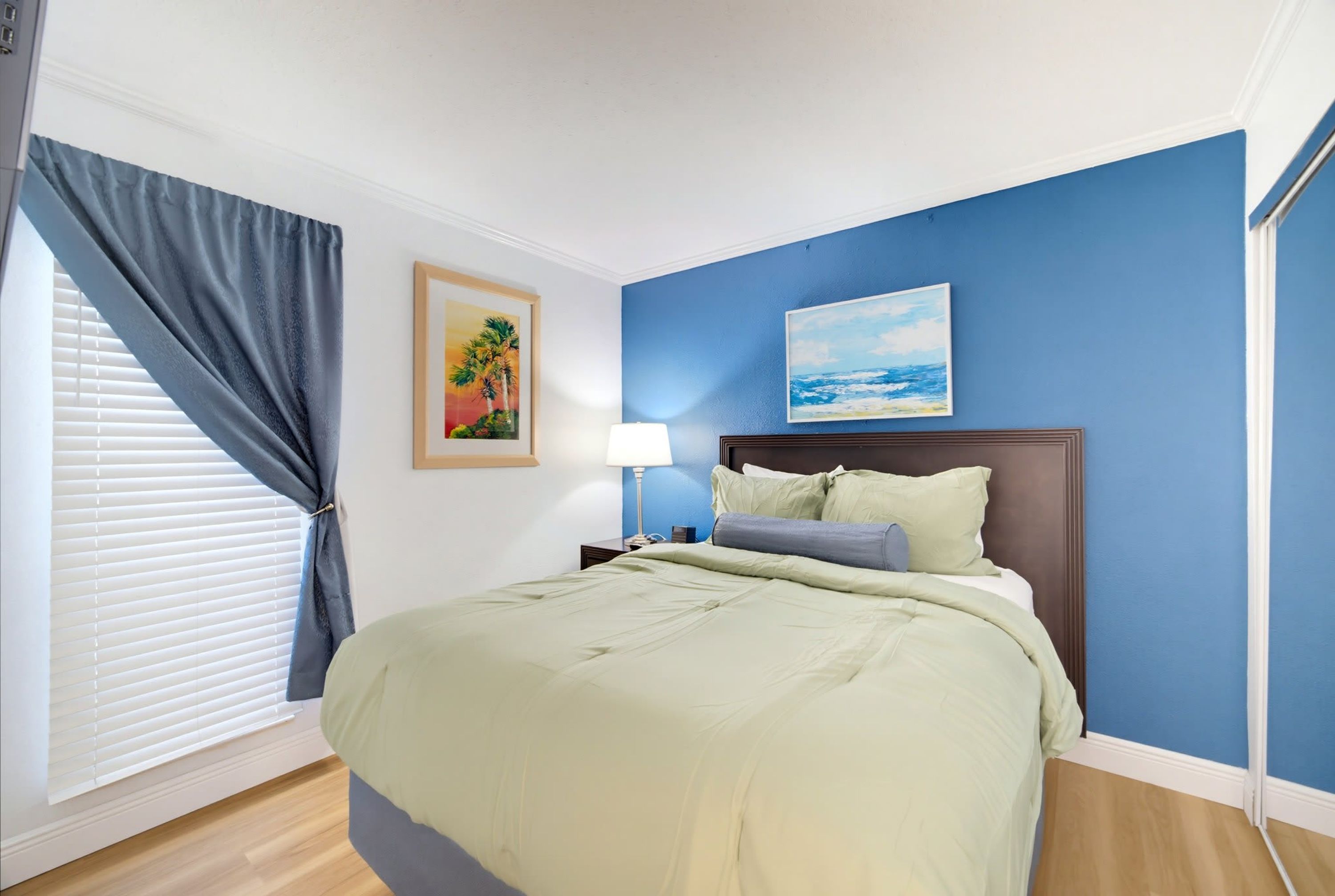A hotel room with a bed, a nightstand, a lamp, a window with blinds, and two framed pictures on the wall.
