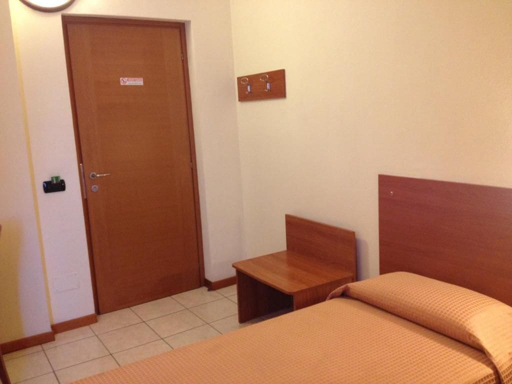 Una habitación con una cama, una mesita de noche de madera, una puerta y un gancho fijado a la pared.