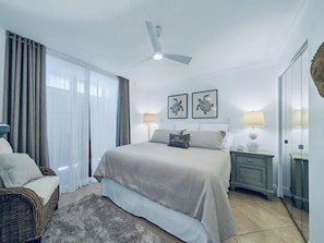 A bedroom with a bed, two bedside tables, a ceiling fan, and a large window with curtains. - Destin Pointe Magnolia House #306 | Luxe Condo, Beautiful Views + Amenities (Destin)