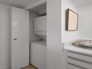A laundry room with a washing machine, a drying rack, and a door leading to another room. - Destin Pointe Magnolia House #306 | Luxe Condo, Beautiful Views + Amenities (Destin)