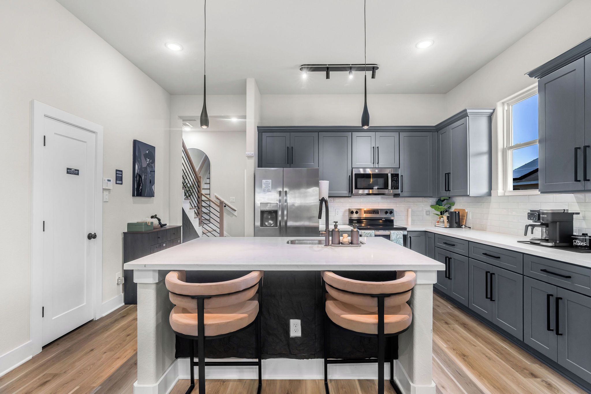 A modern kitchen with a central island, grey cabinets, stainless steel appliances, and a staircase leading to another level.