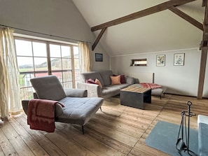Ein Loft-Zimmer mit Holzboden, einem grauen Sofa, einem grauen Sessel und einem kleinen Holztisch. Ein Fenster mit Vorhängen, und ein Balkon ist durch das Fenster zu sehen.