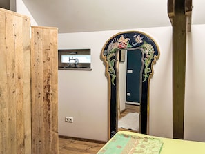 Ein Zimmer mit einem Holzschrank, einem Spiegel mit aufwendigen Verzierungen, einem kleinen Fenster und einem Bett mit einem gemusterten Überwurf.
