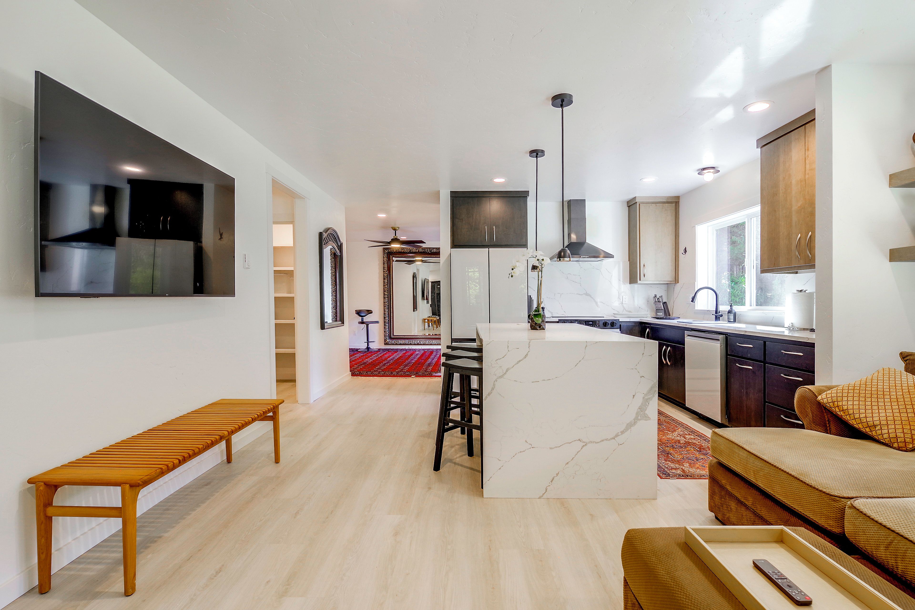 Una cocina moderna con una isla central, un comedor con banco y una sala de estar con sofá y televisor.