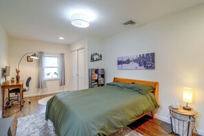 Un dormitorio con cama, una mesita de noche con lámpara, una estantería, una ventana con cortinas y un cuadro colgado en la pared.