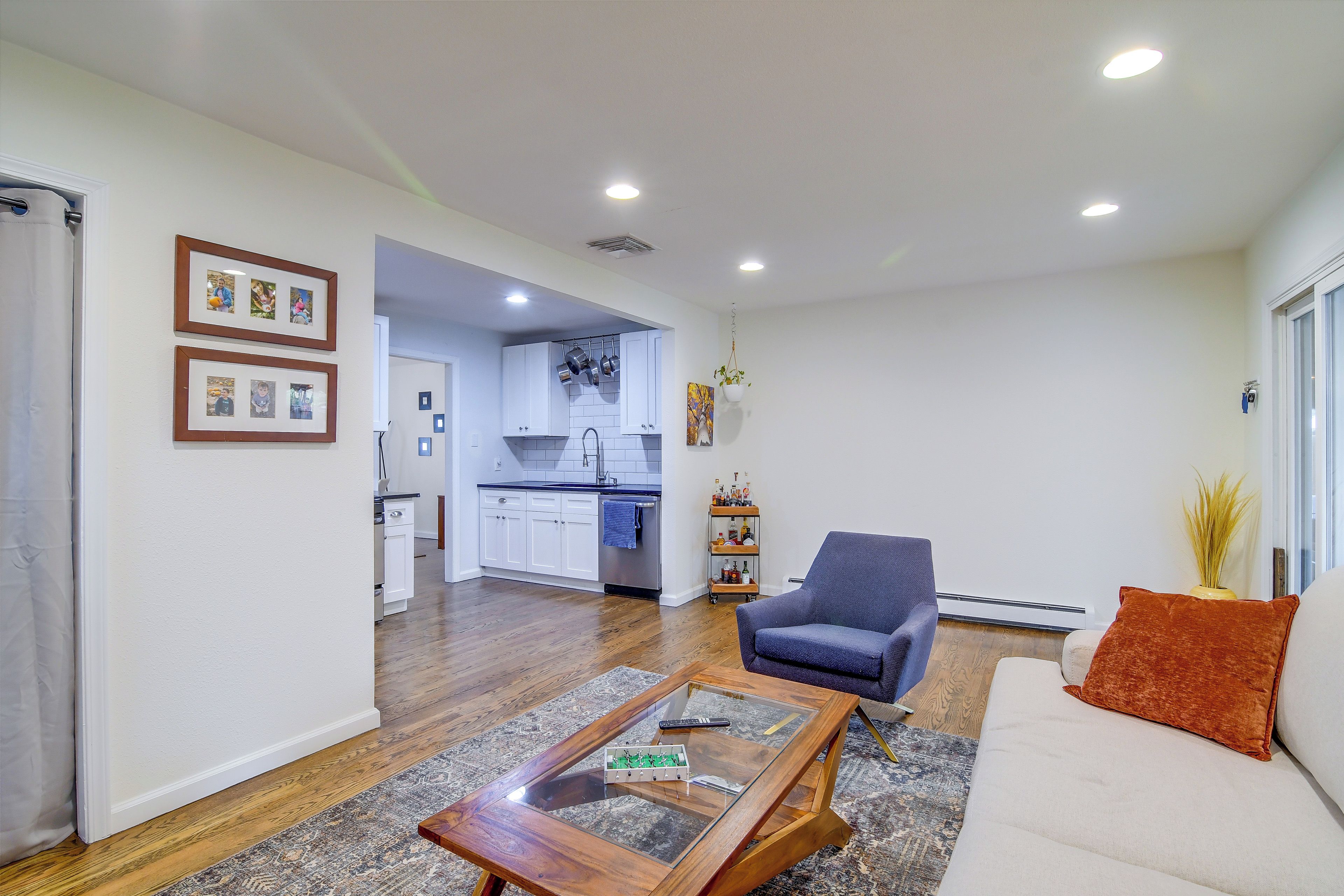 A living room with a sofa, a coffee table, and a chair. There is a kitchen in the background with white cabinets and a hanging pot rack.