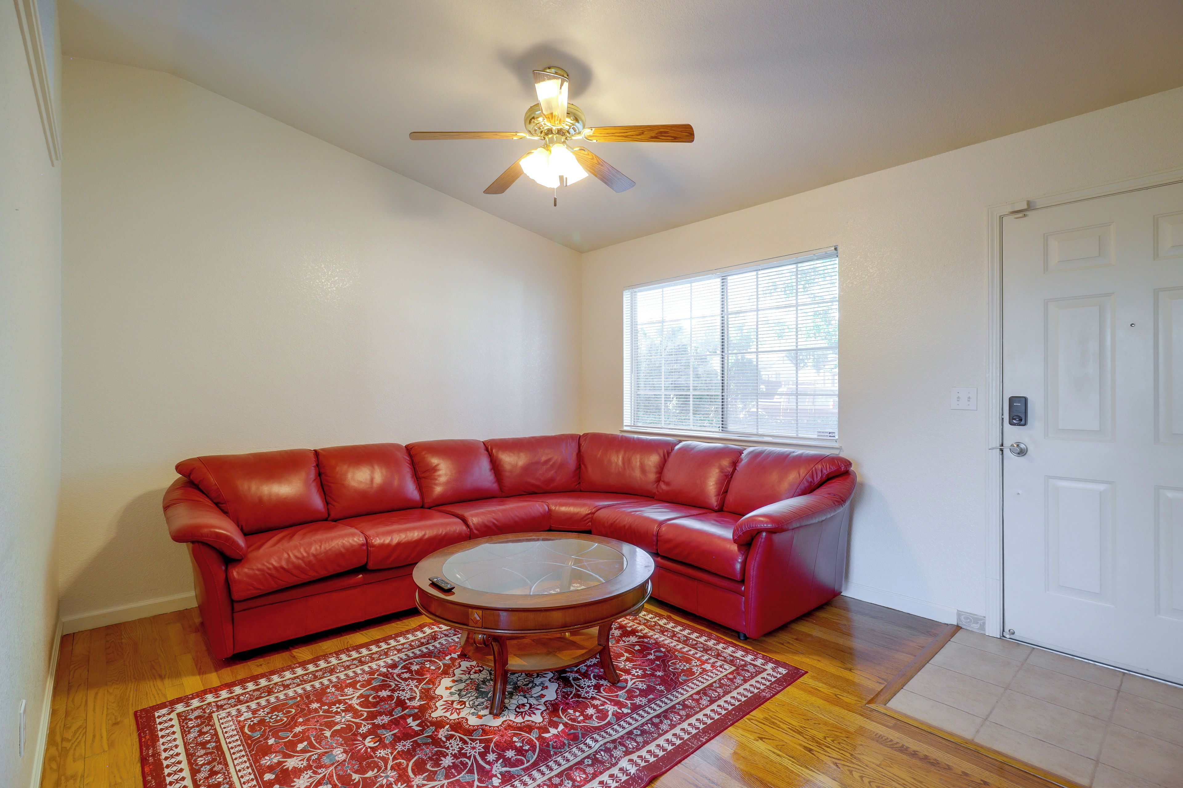 A living room with a red leather sofa, a glass coffee table, a patterned rug, a ceiling fan, and a window with blinds.