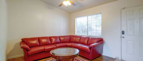 A living room with a red leather sofa, a glass coffee table, a patterned rug, a ceiling fan, and a window with blinds.