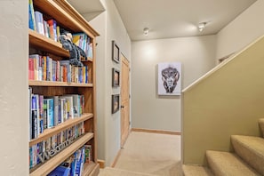 A hallway with wooden bookshelves filled with books, a framed picture on the wall, and a carpeted staircase. - Moonlit Hearth & Hickory | Ski-In/Out + Hot Tub (Big Sky)
