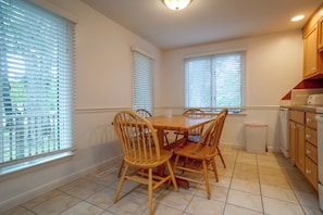 A dining area with a wooden table and chairs, a kitchen with wooden cabinets, and windows with blinds. - Retreat 2 Mi to Cape Cod Beaches! Bring Your Pets (Mashpee)