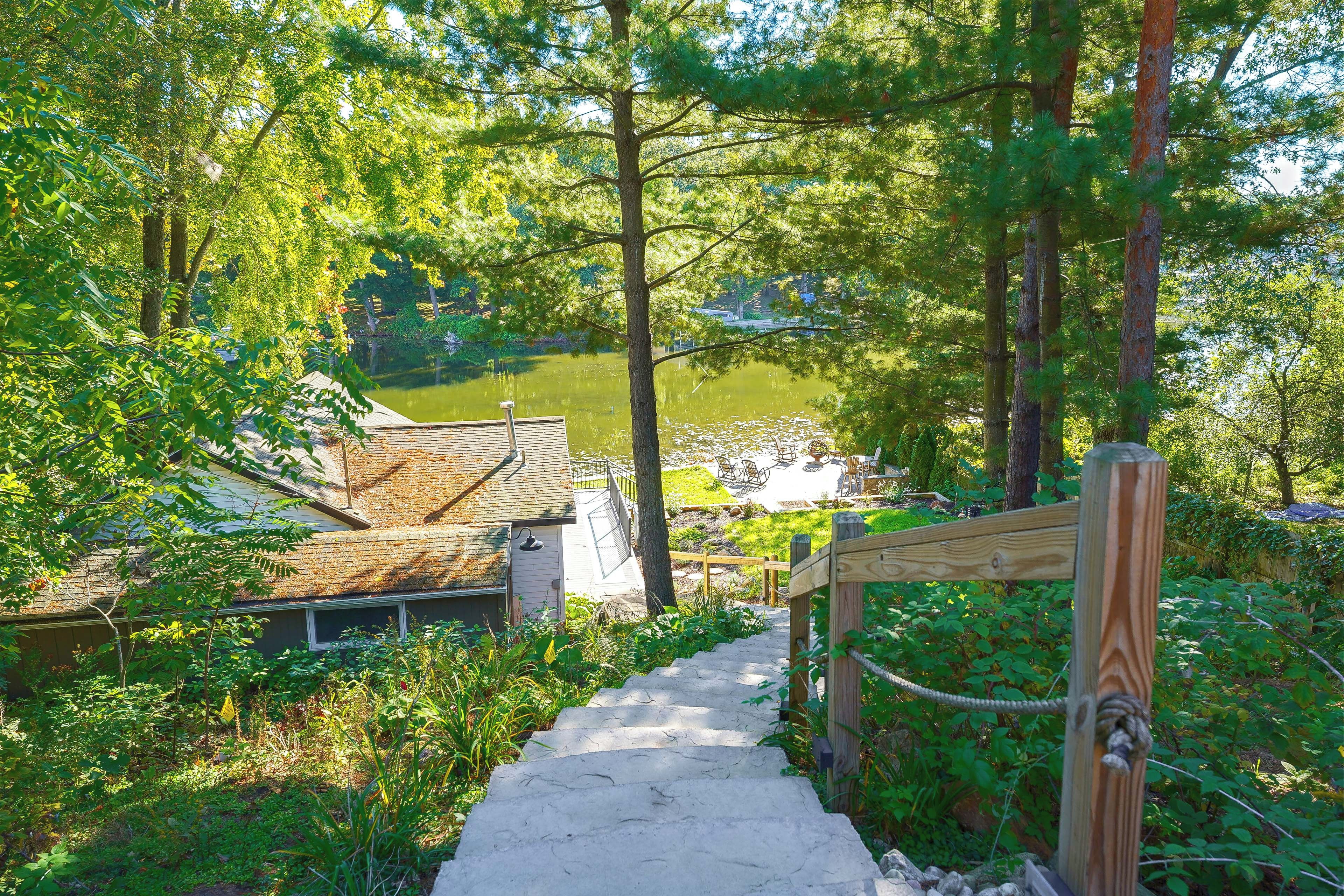 A wooden pathway leading to a building with a sloped roof, surrounded by lush greenery and a body of water.