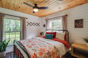 A bedroom with a bed, a ceiling fan, a window with curtains, a wall-mounted decoration, and a potted plant.