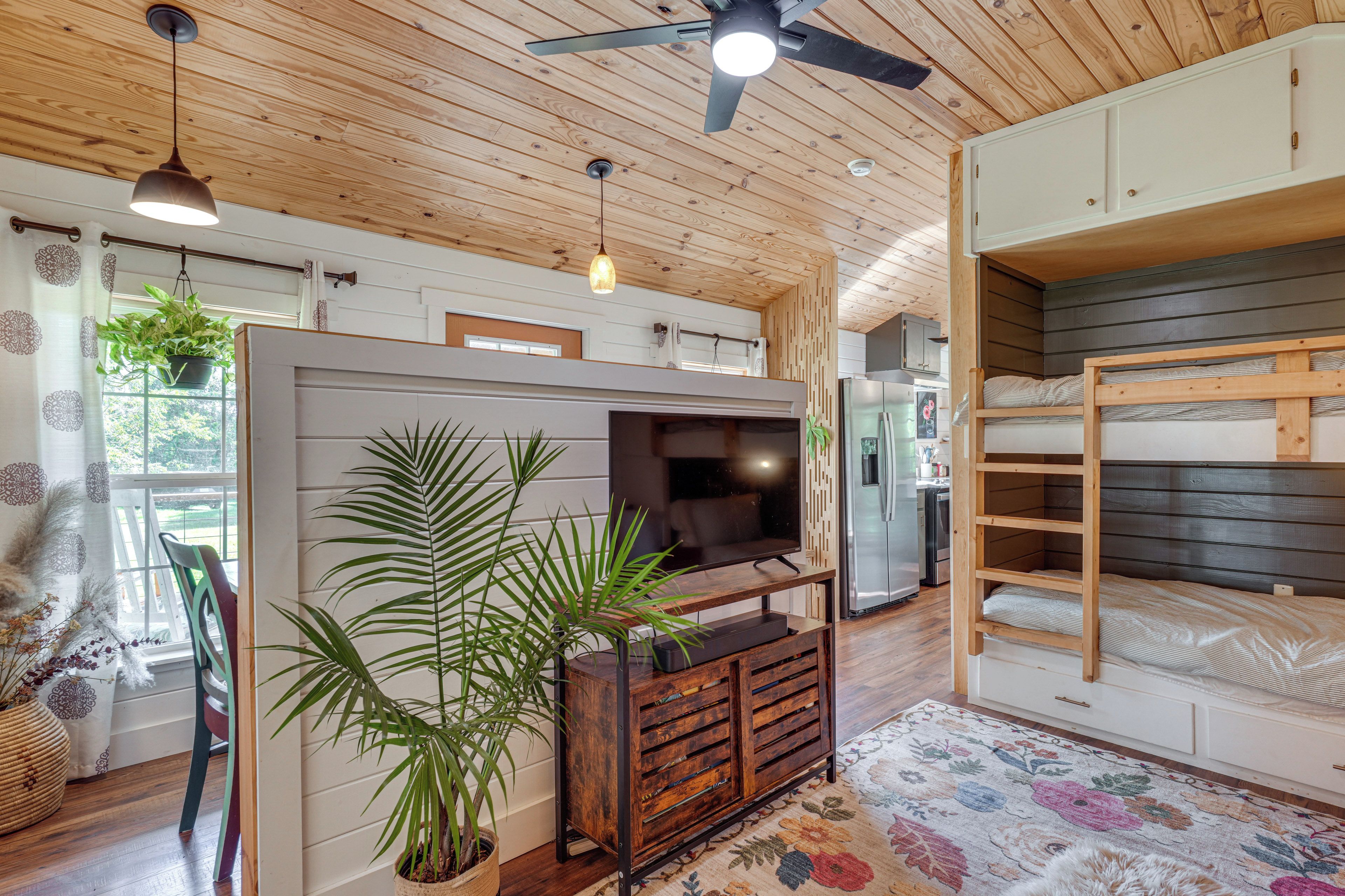 A room with a wooden ceiling, a ceiling fan, a television on a wooden stand, a bunk bed, and a potted plant.