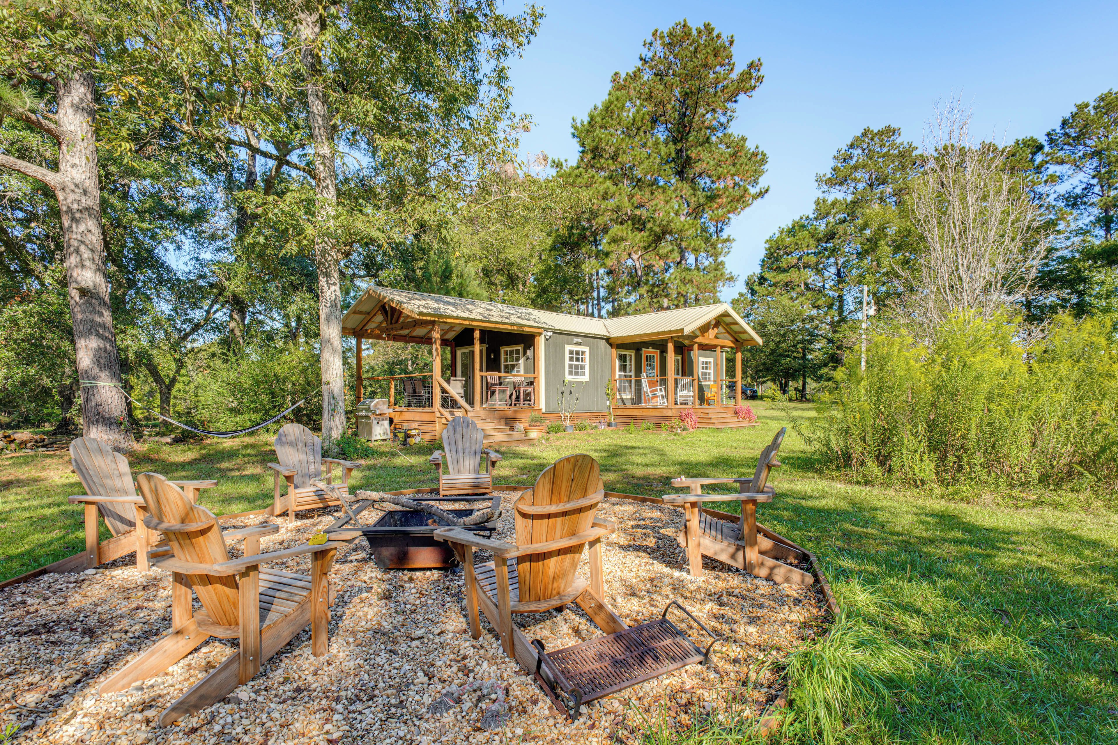A cabin with a deck, surrounded by trees, and a fire pit with Adirondack chairs.