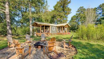 A cabin with a deck, surrounded by trees, and a fire pit with Adirondack chairs.