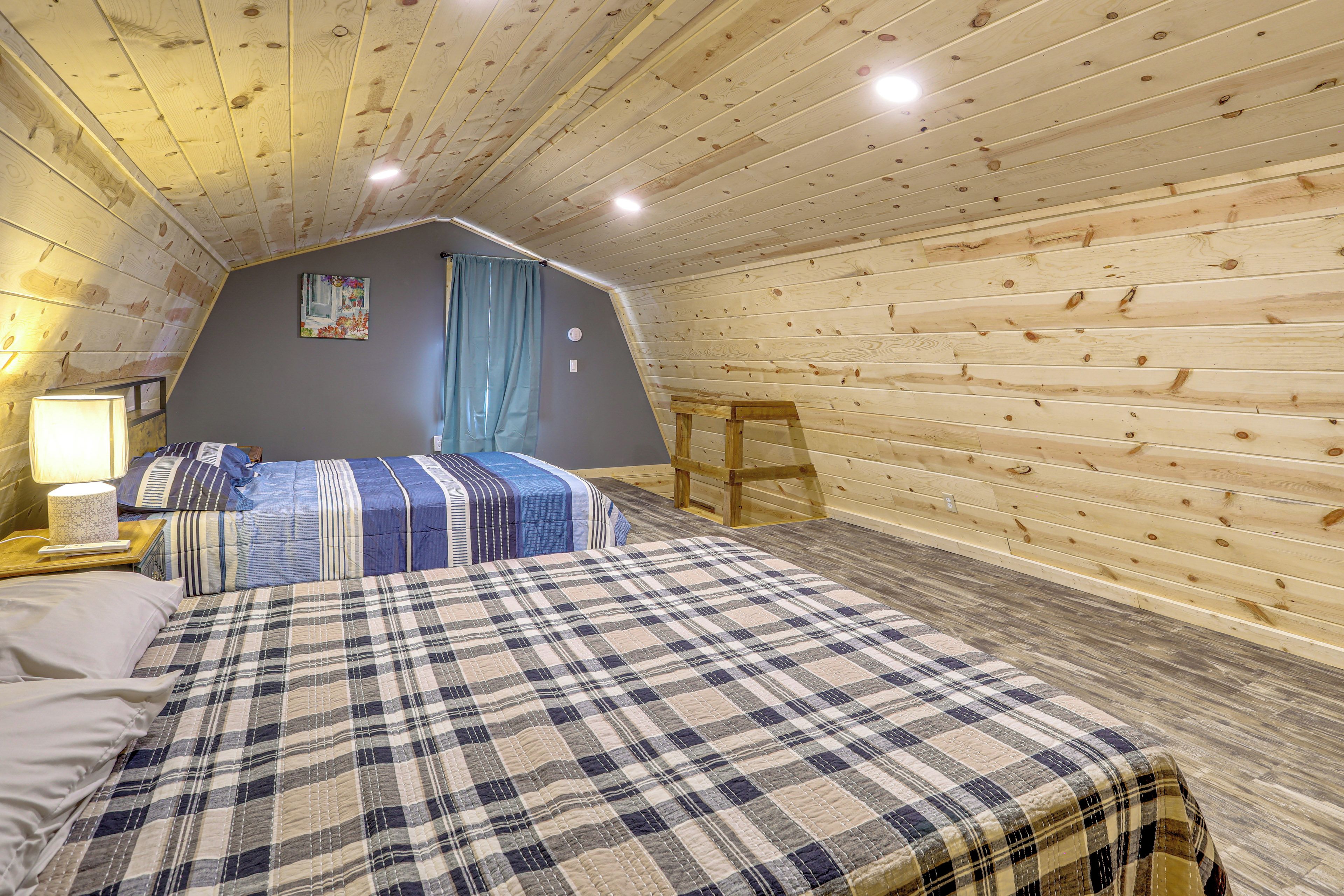 A bedroom with a sloped ceiling, a bed with a plaid bedspread, a nightstand with a lamp, and a window with curtains.